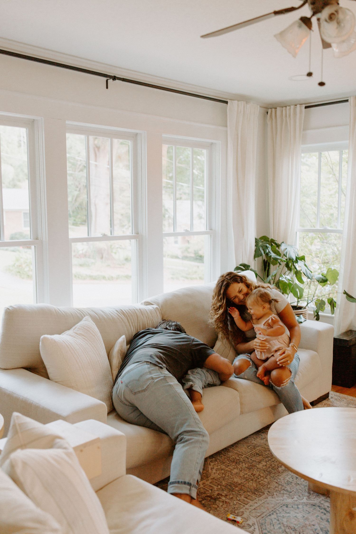 Candid moments of family playtime on a plush white couch with natural light streaming through windows.