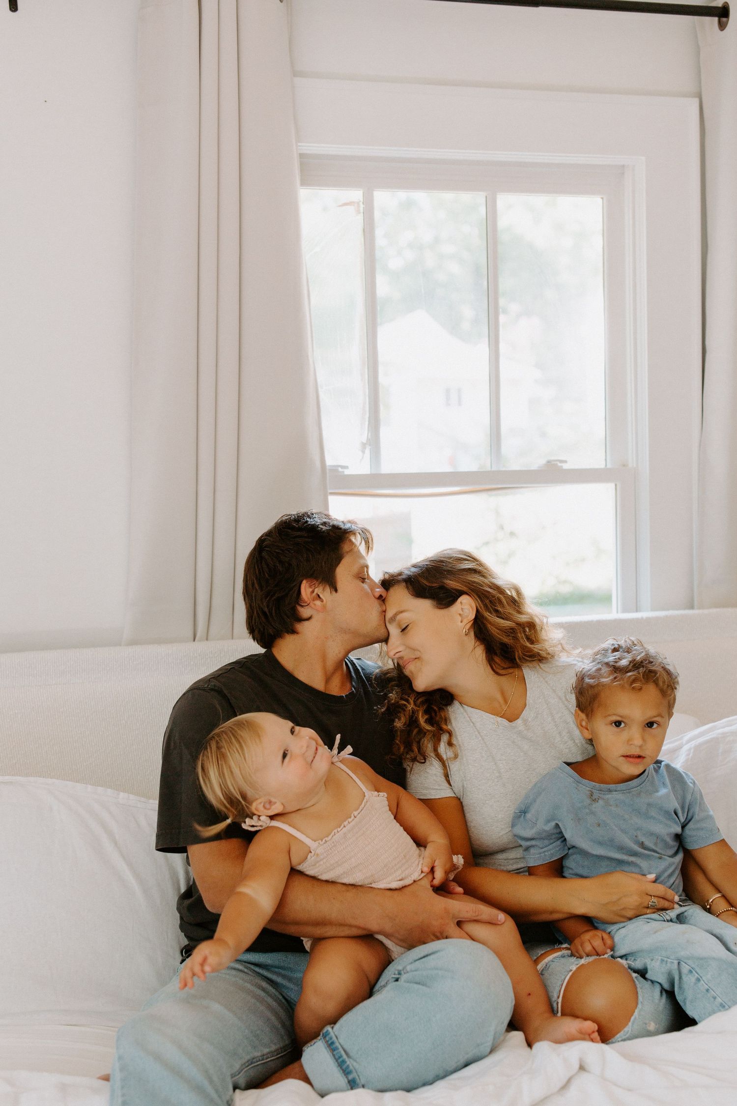 An intimate family moment shared on a bright white bed near a window.