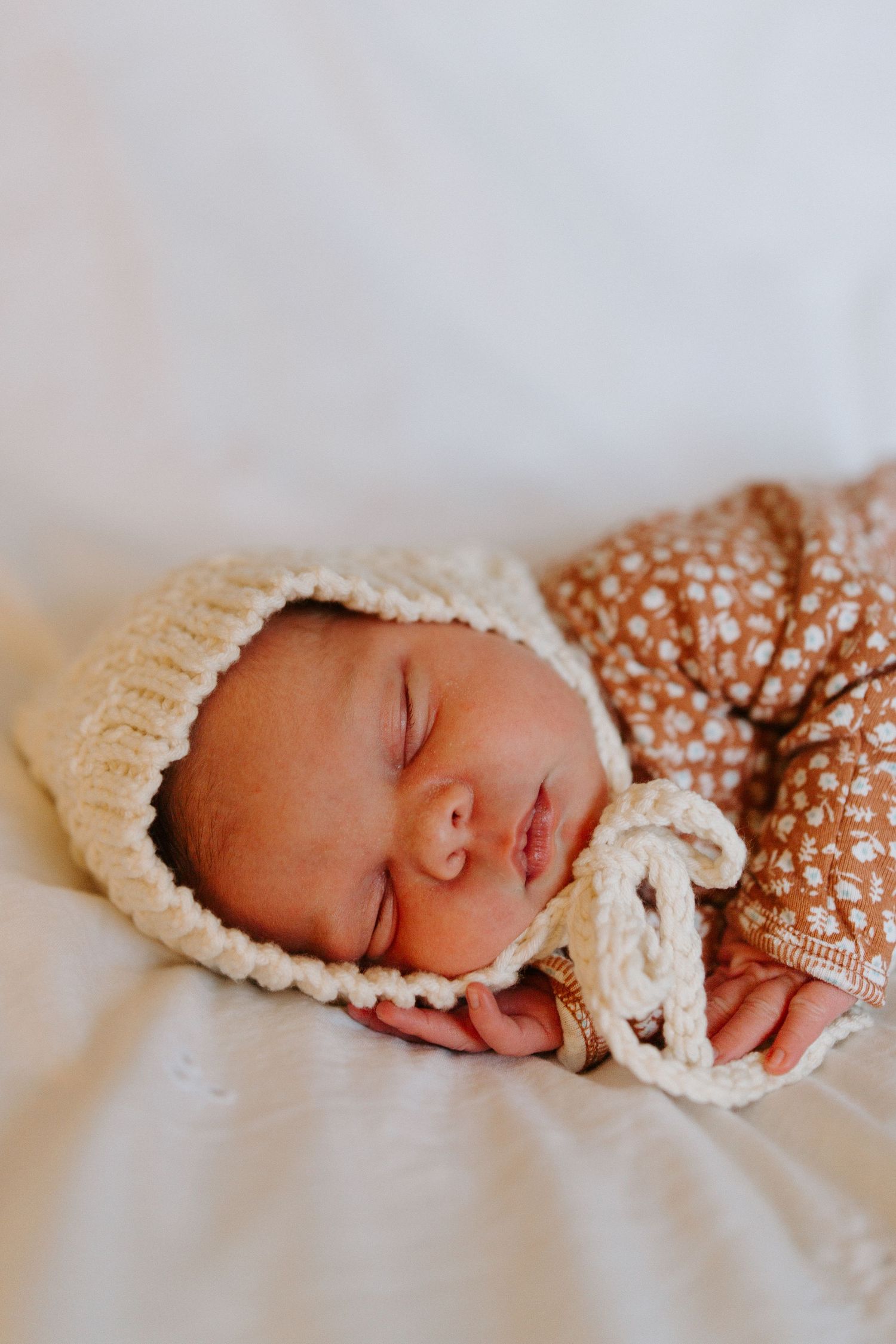 A sleeping newborn baby wearing a knit bonnet rests peacefully on white bedding.