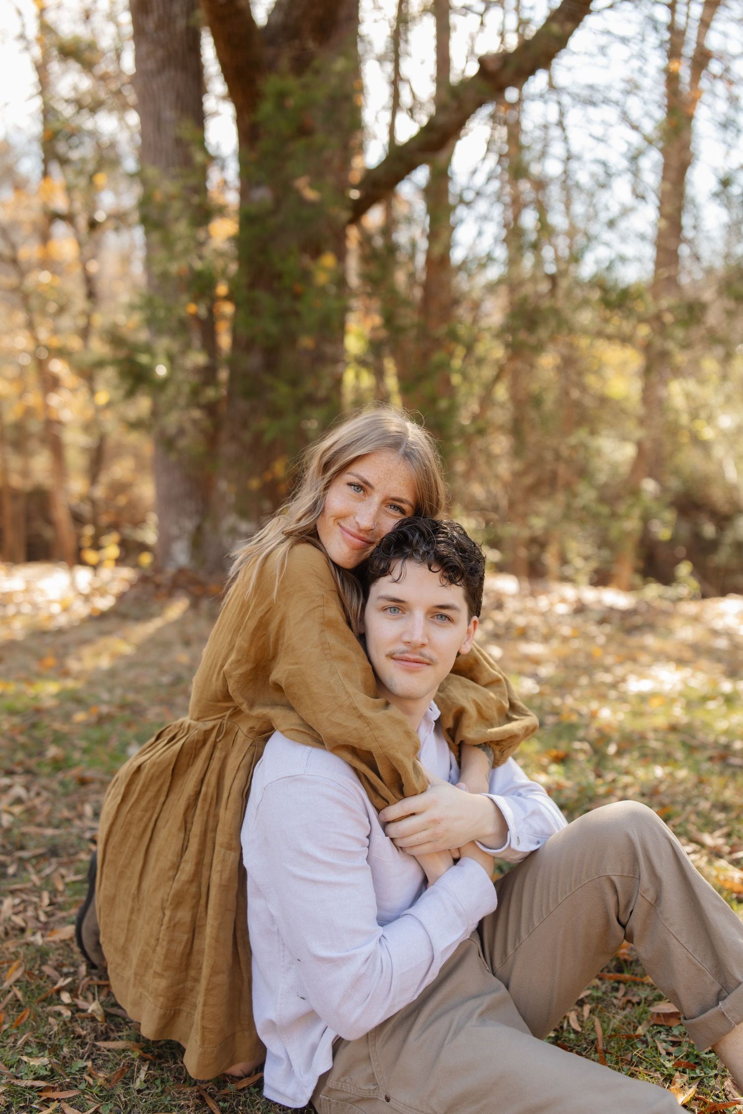 A young couple embraces outdoors in autumn, woman in mustard dress hugging man from behind near trees.