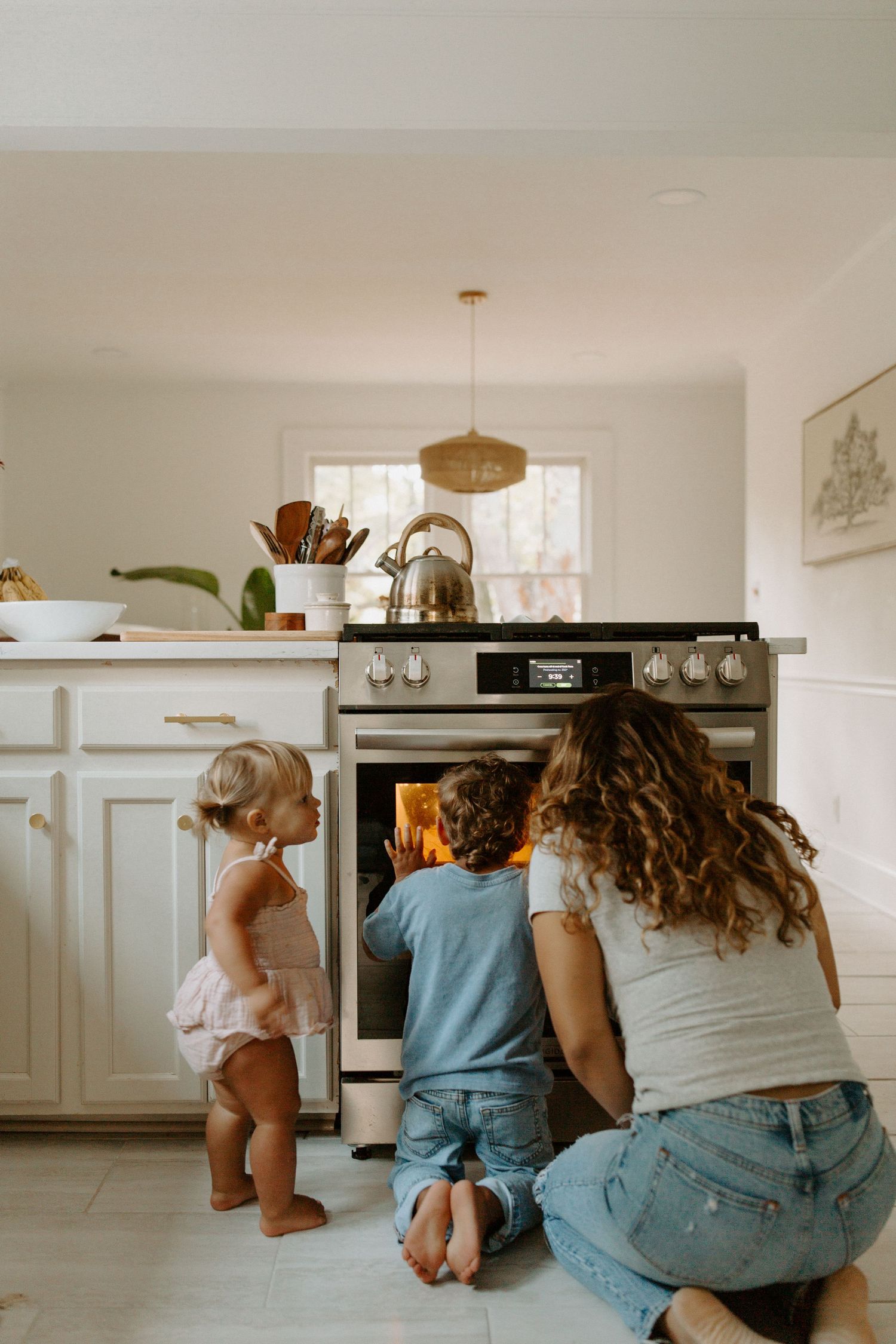 Three people kneeling to peek into illuminated oven in white kitchen.