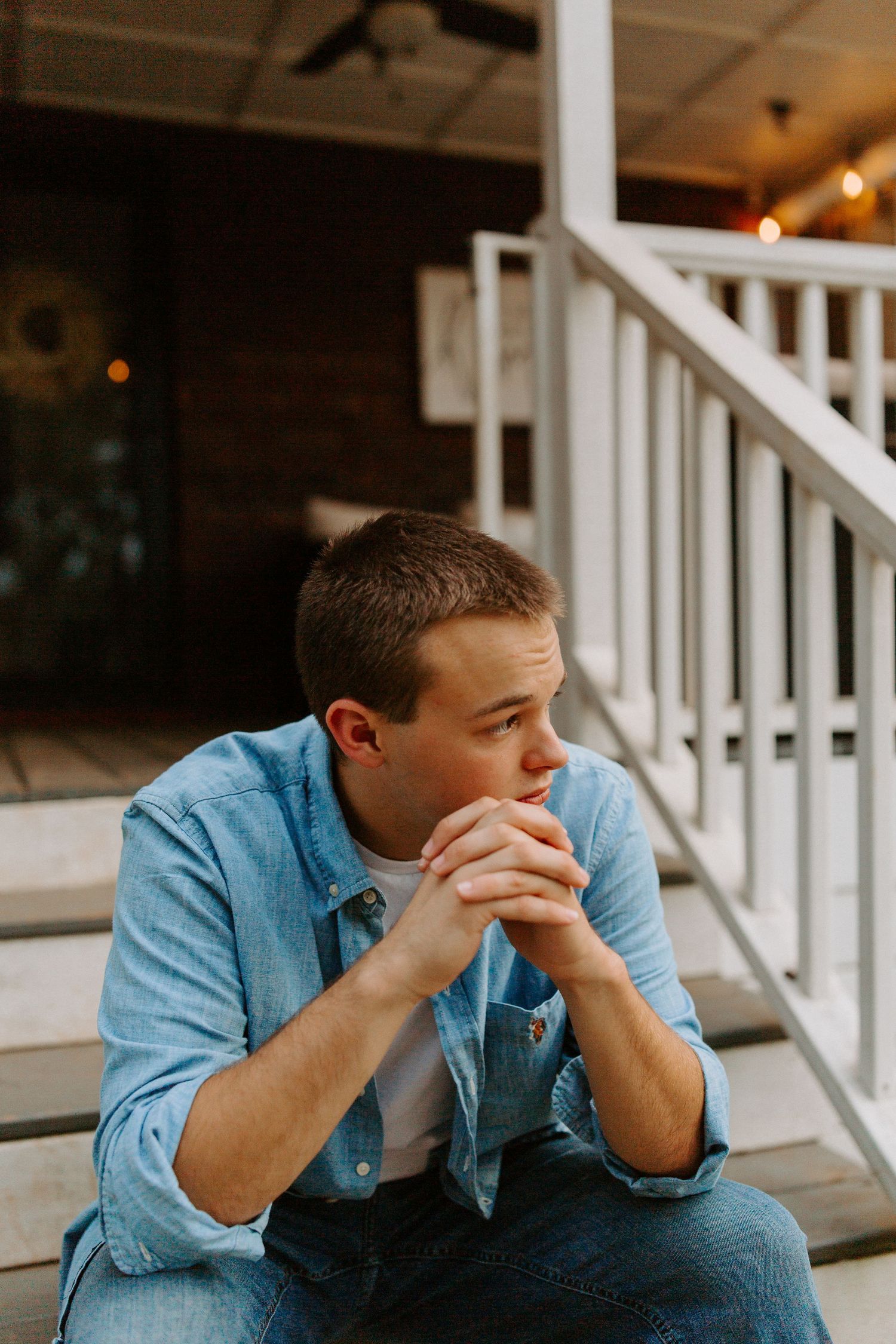 Person in denim jacket sits thoughtfully on white porch railing at dusk.