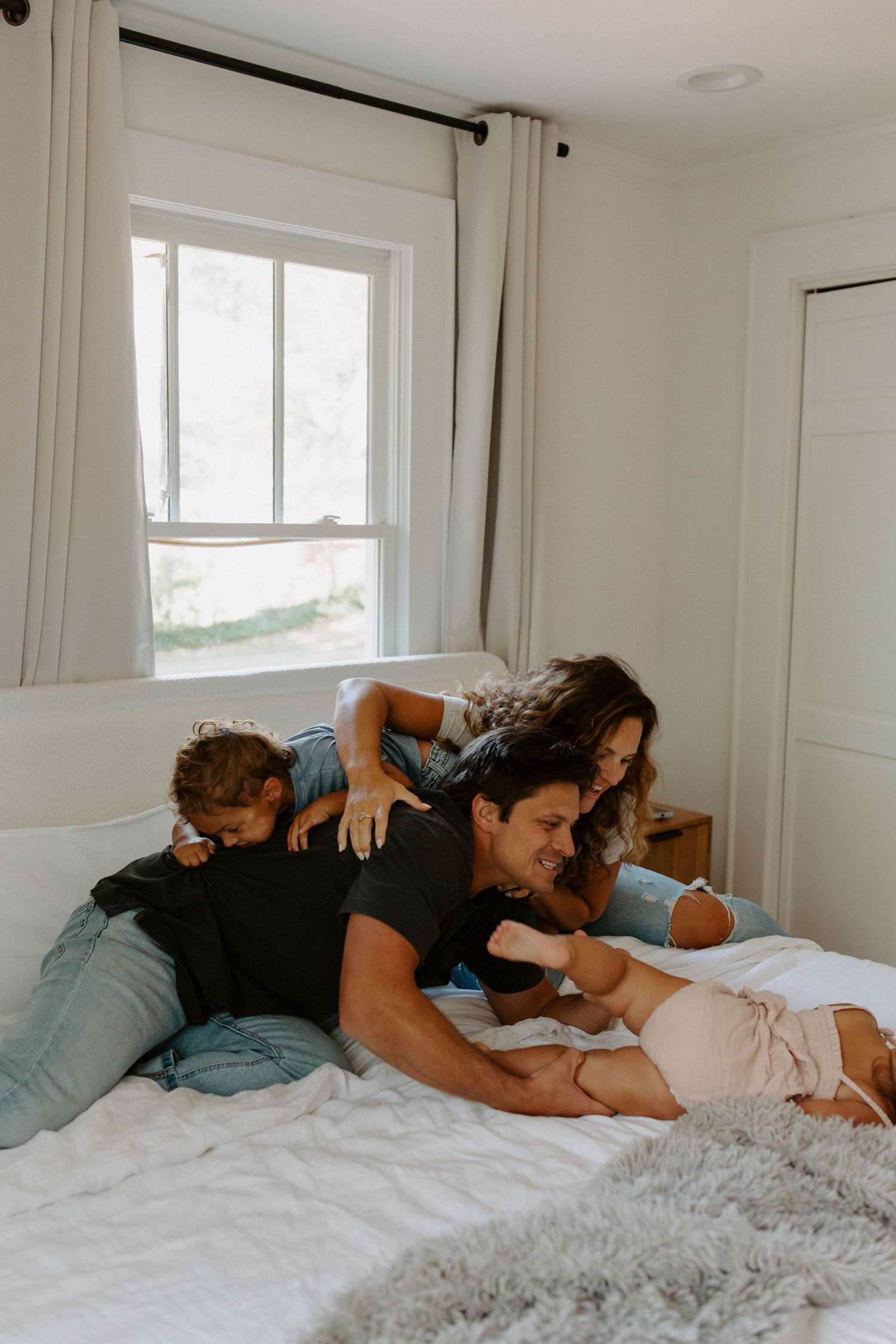 A family lounges together on a white bed in a bright bedroom with natural light streaming through the window.