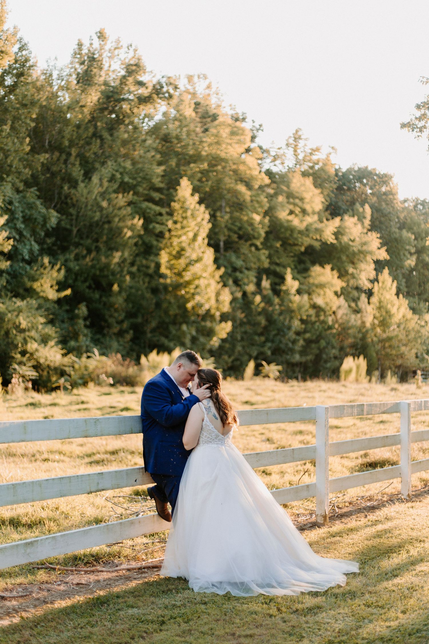 Newlywed couple shares intimate moment by white fence at sunset.
