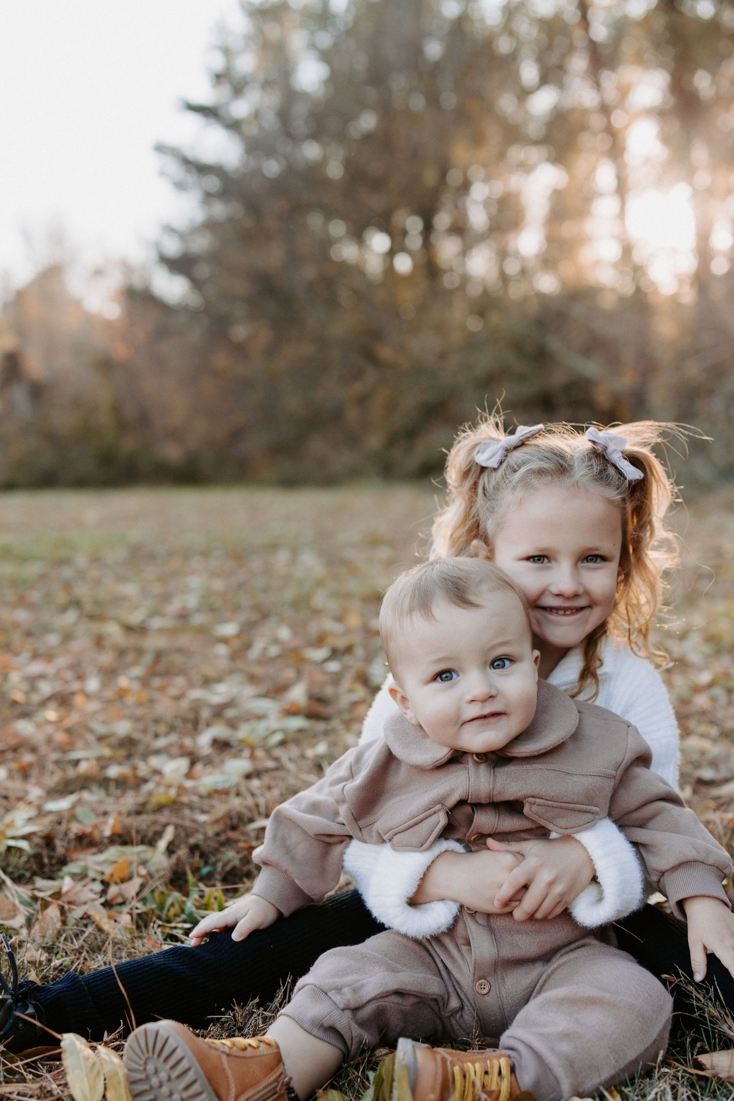 Children sitting together among fallen autumn leaves in matching beige outfits.