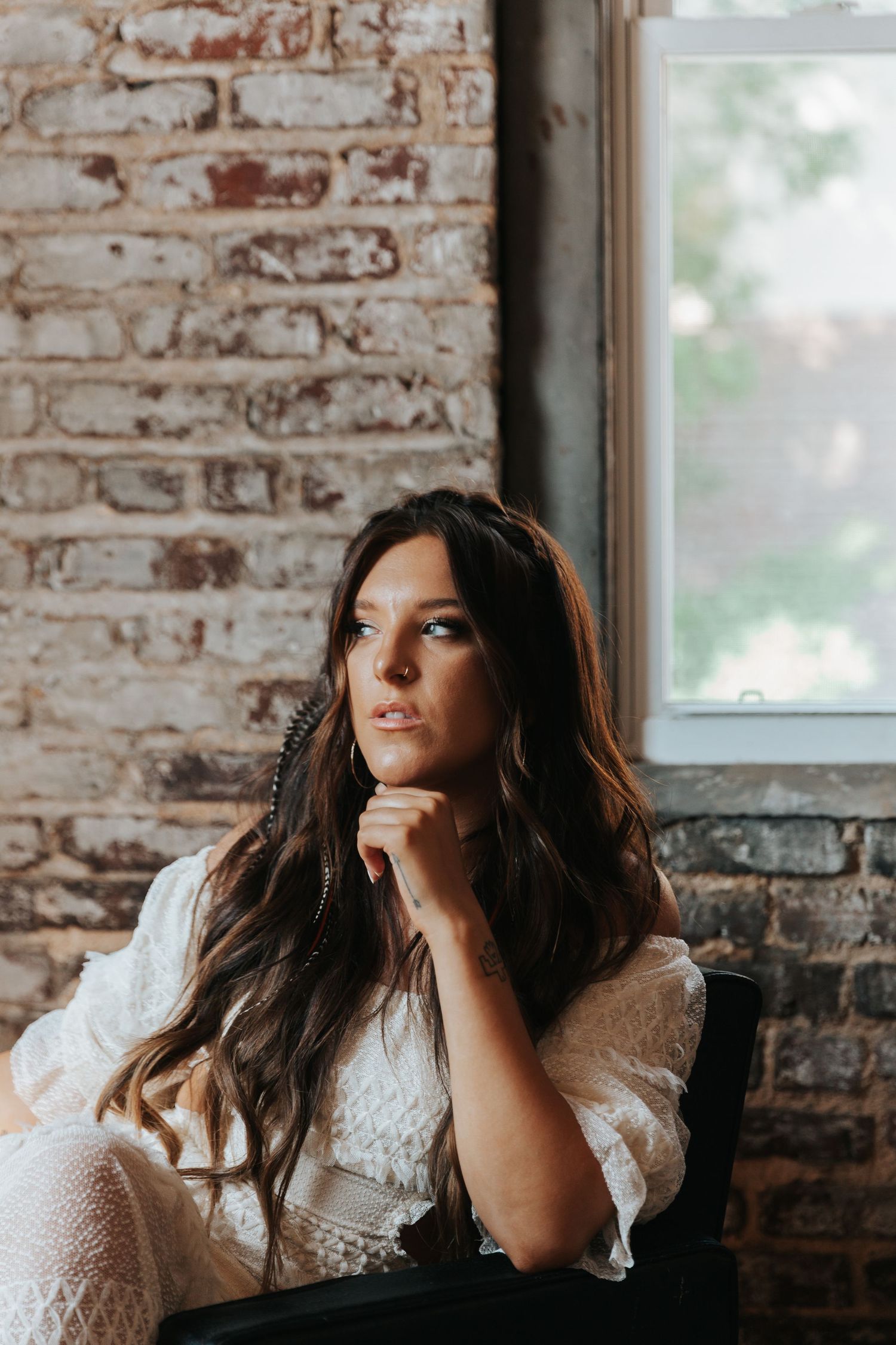 Portrait of woman in white lace dress seated by window with exposed brick wall.