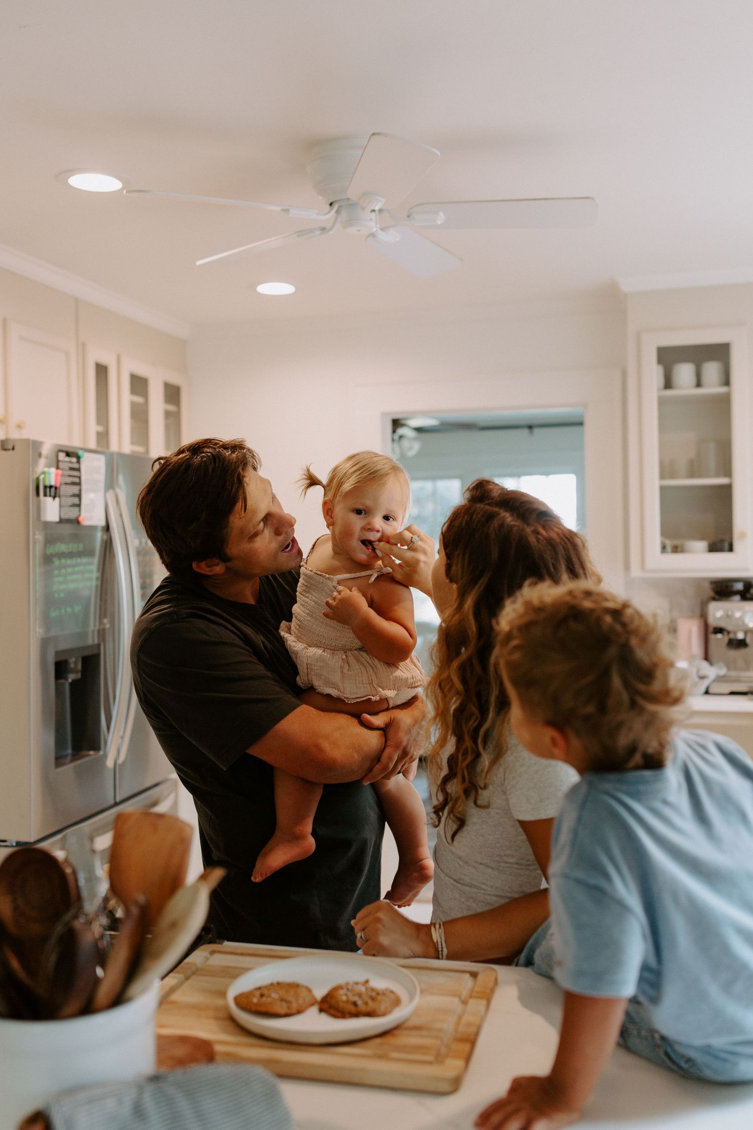 A joyful family moment in a bright white kitchen with cookies on a wooden cutting board.