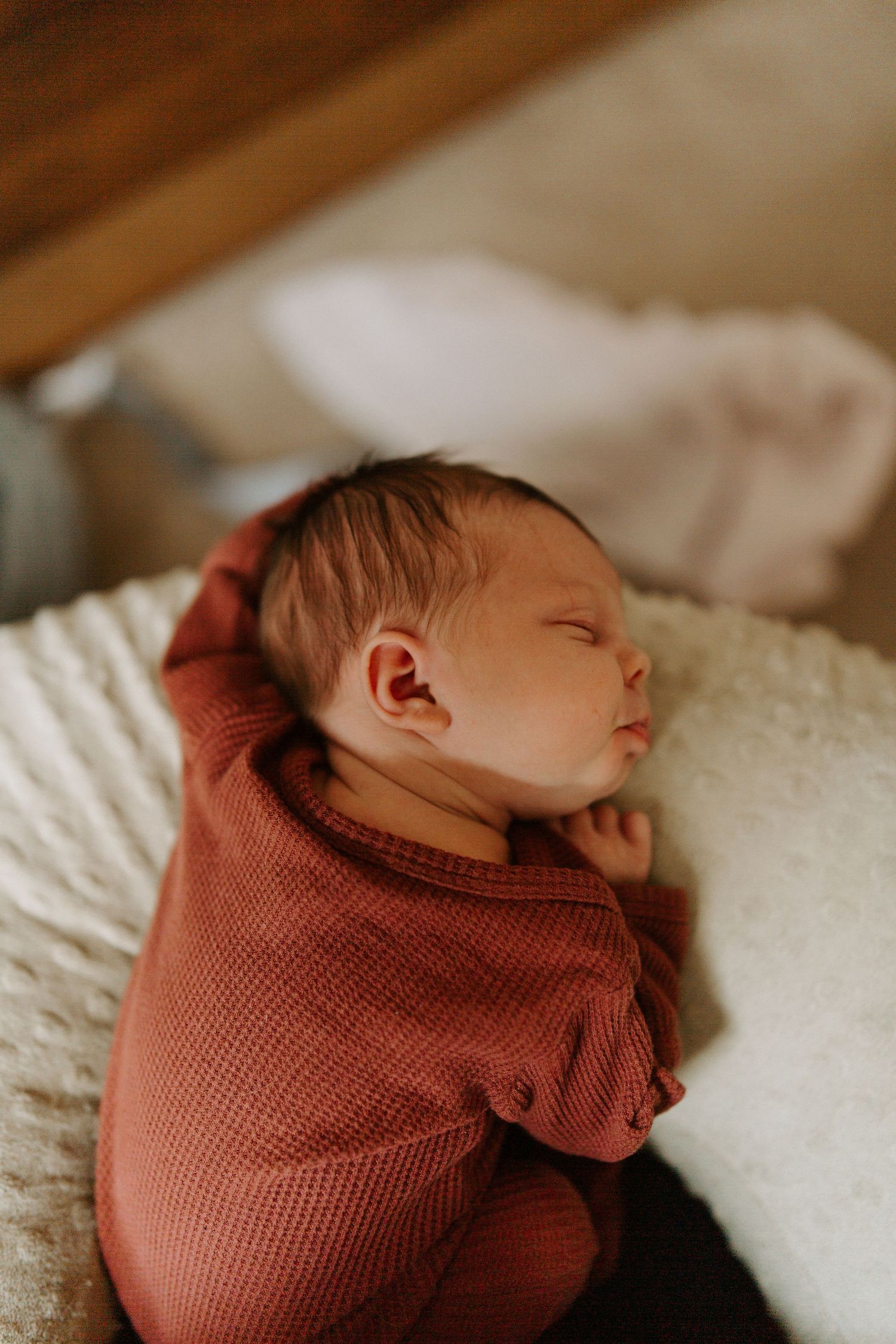 Newborn baby wrapped in rust-colored knit swaddle resting on white blanket.