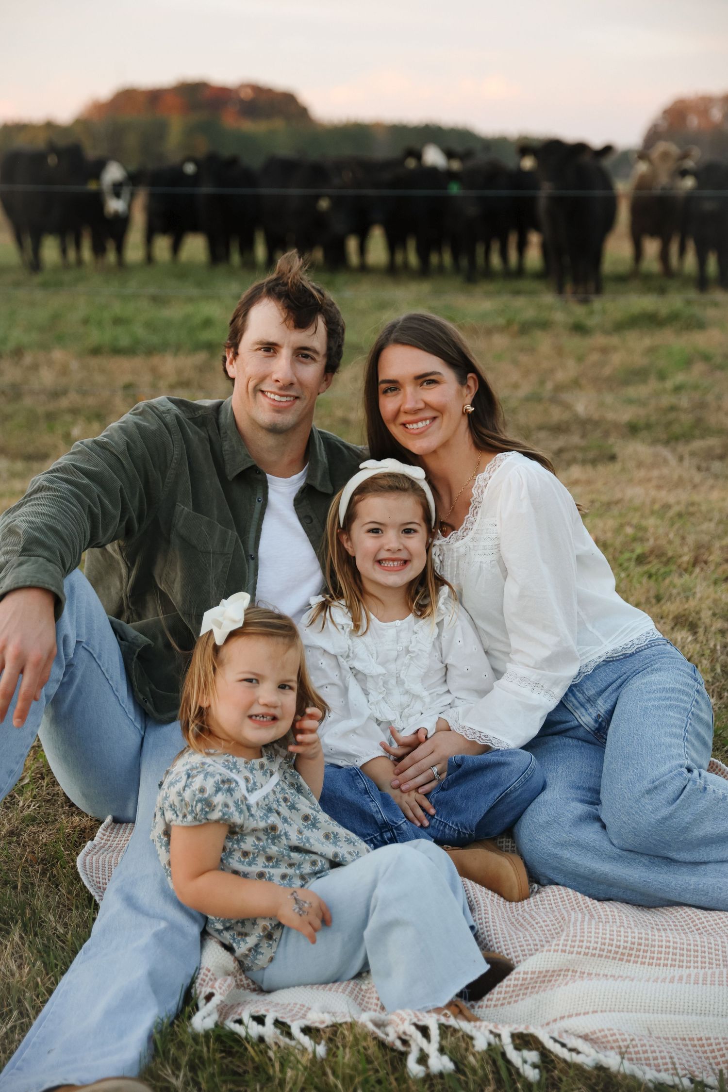 Happy family of four sitting on a blanket in a field with black cattle grazing in the background.