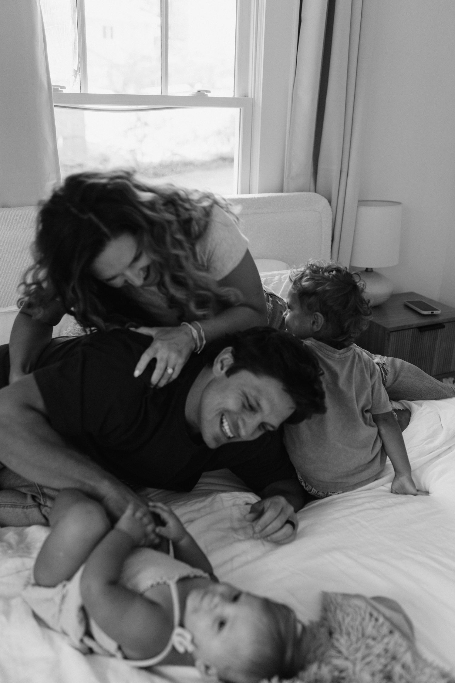 A candid black and white photo of a family playfully lounging together on a bed near a window.