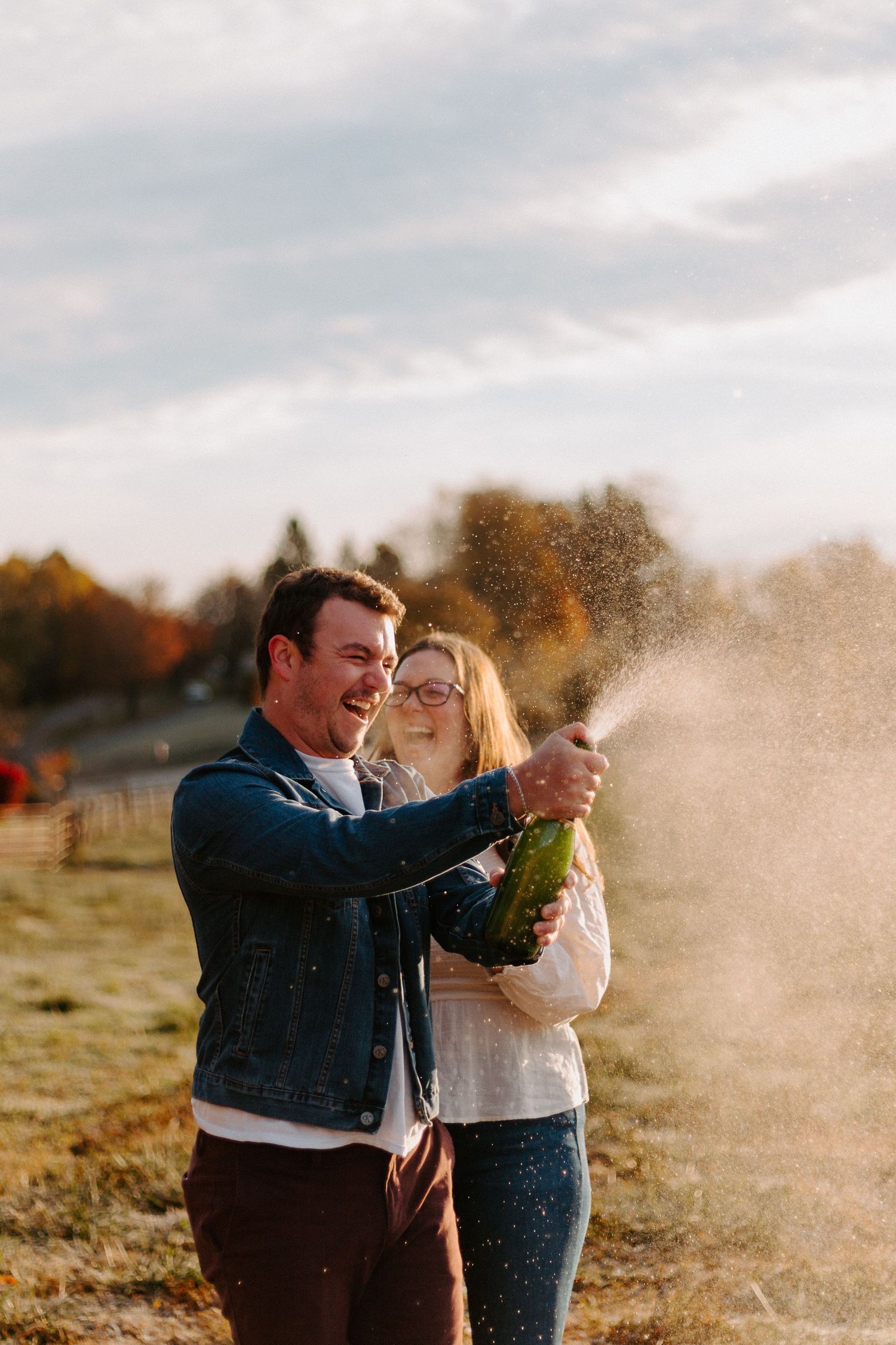 A couple celebrates with champagne spraying during a golden hour photoshoot in an autumn field.