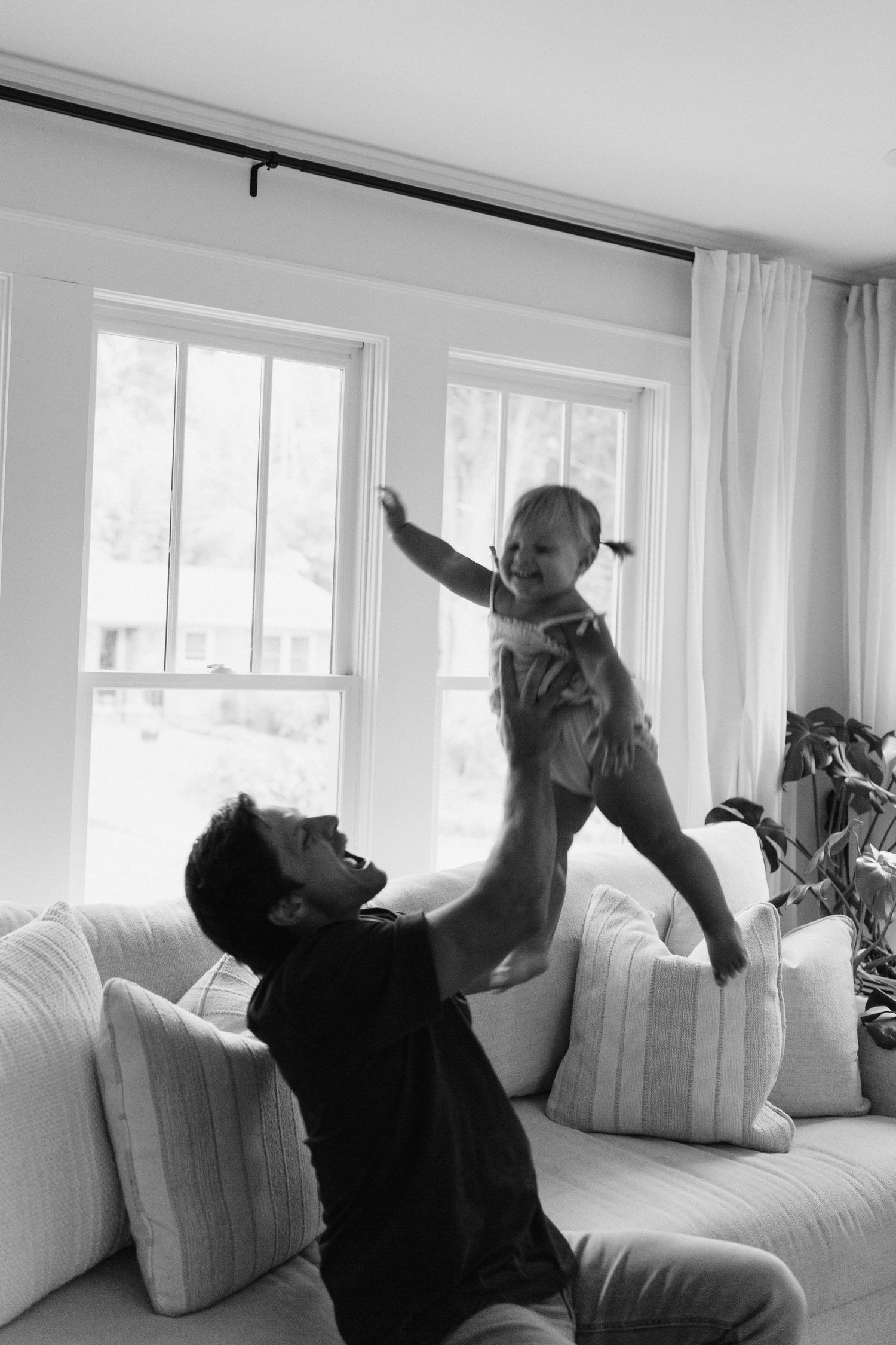 Playful family moment on a white couch in a bright sunlit room with large windows.