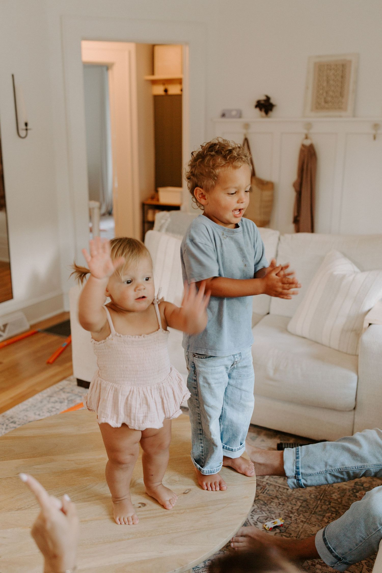 Toddlers in casual clothing playing and dancing on hardwood floors near white sofa.