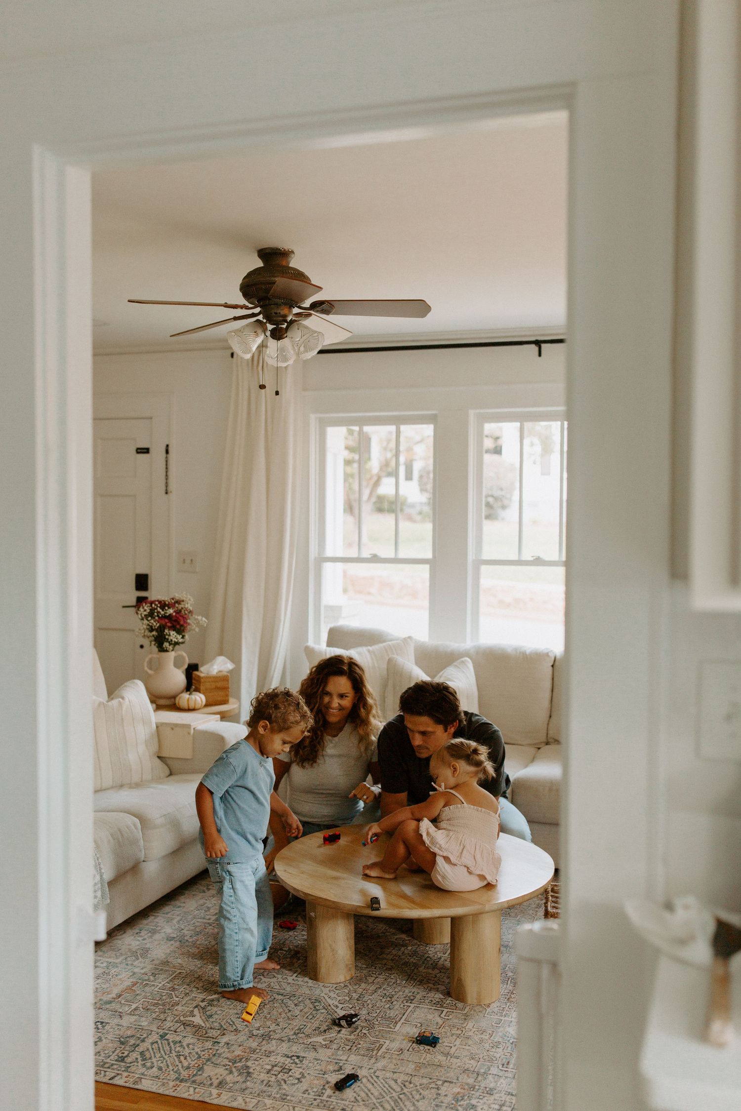 Family gathered around wooden coffee table in bright living room with ceiling fan.