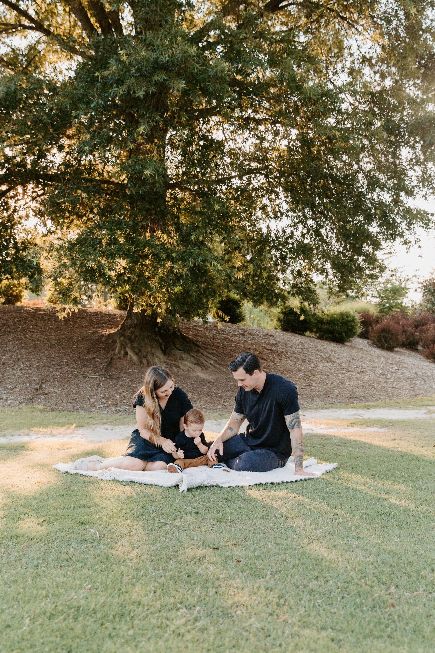 Family relaxing on blanket under large tree during outdoor photoshoot.