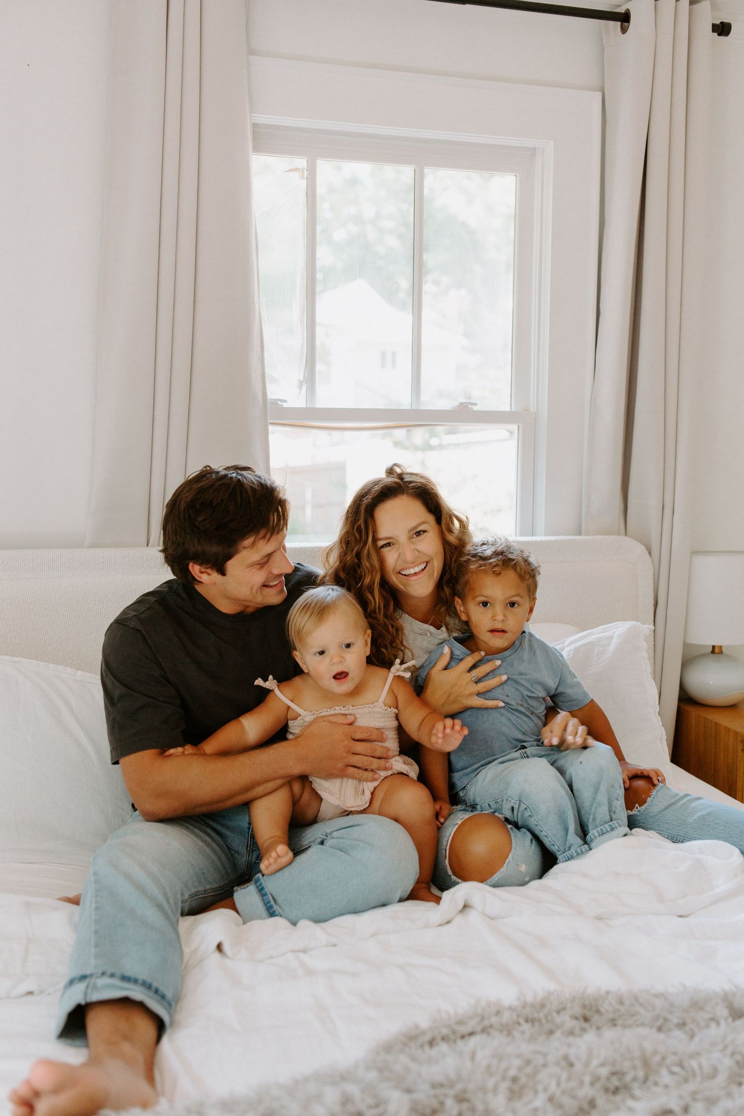 A loving family moment captured on a cozy white bed in a sunlit room.