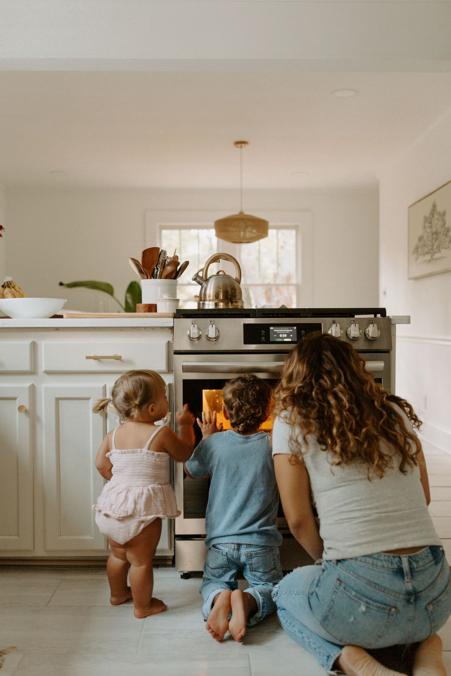 Family gathered around oven watching something bake in bright modern white kitchen.