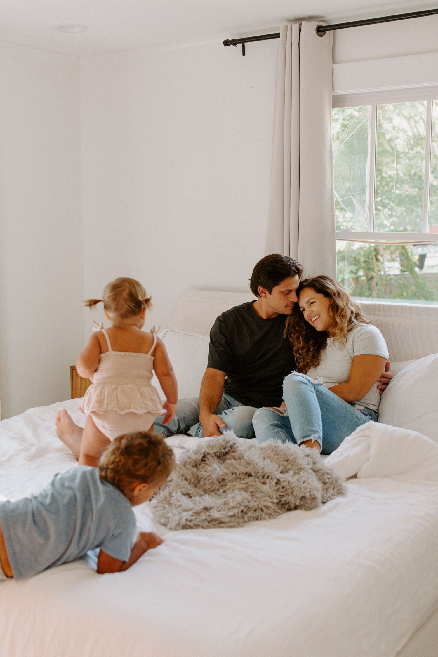 Family relaxing together on white bedding in bright, airy bedroom.