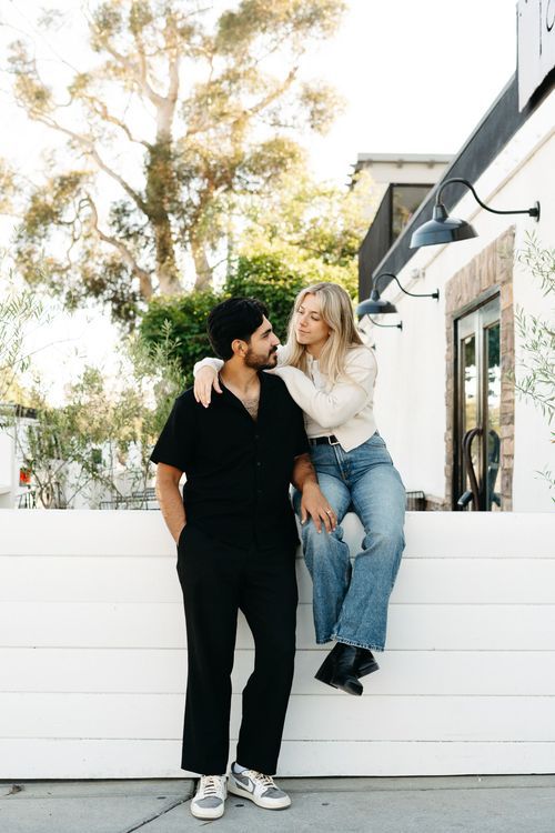 Couple sitting and interacting playfully against white wall outdoors.