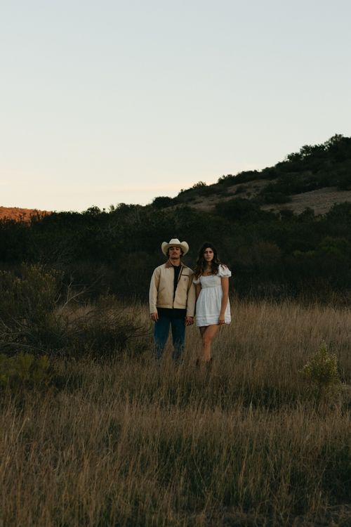 Couple holding hands in a field wearing white dress and cowboy hat.