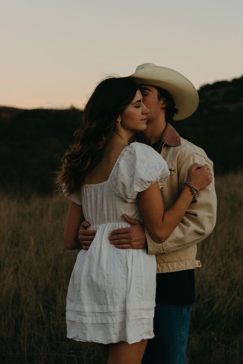 Romantic couple embracing in a golden field at sunset wearing white dress and cowboy hat.
