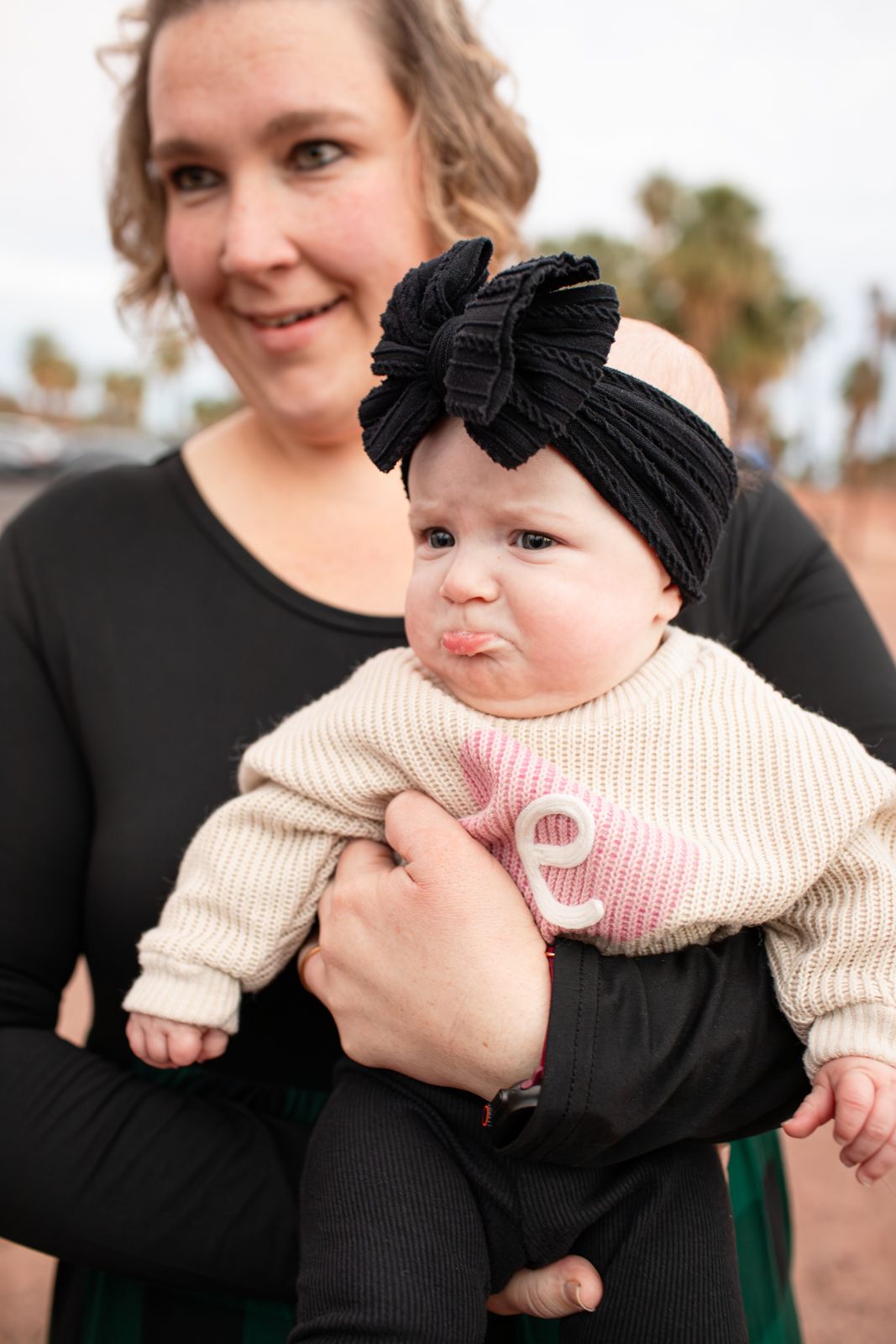 A baby wearing a black bow headband and cream sweater being held outdoors with palm trees in background in Papago Park in Phoenix, AZ.
