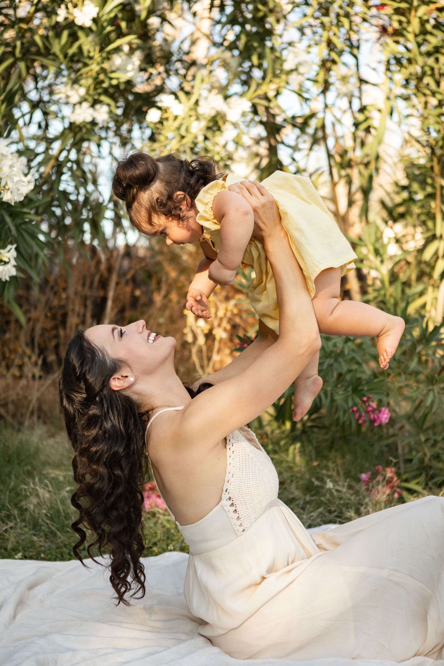 A tender moment captured outdoors as a woman in a cream dress lifts a small child against a sunlit garden backdrop at The Meadow in Mesa.