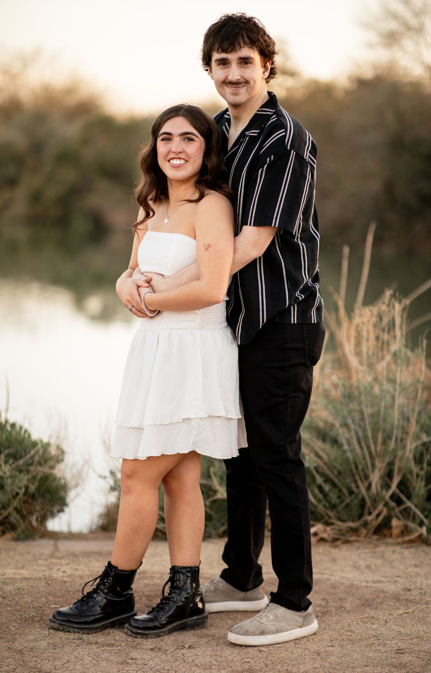 A couple poses together outdoors at sunset, wearing a white dress and black striped shirt by a lake.