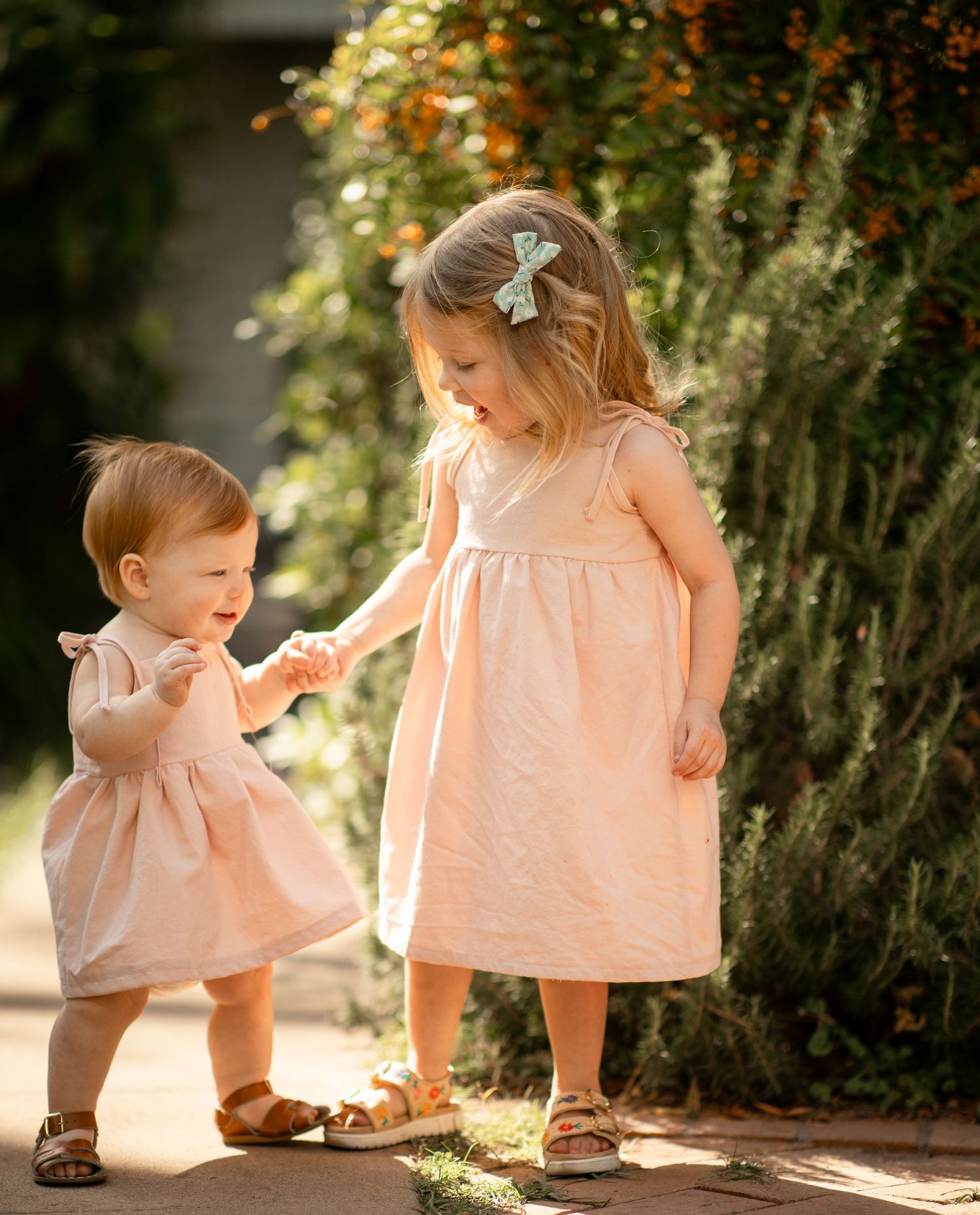 Two young girls in matching pink dresses hold hands outdoors, with lush green foliage in the background.