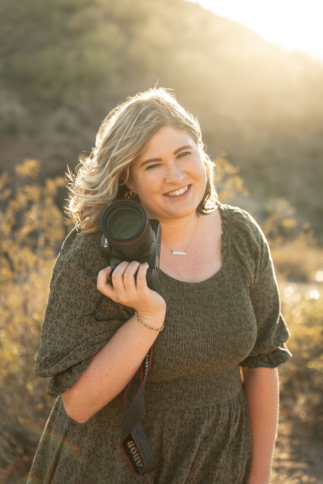 Professional photographer in an olive dress holding a camera during golden hour in a desert landscape.