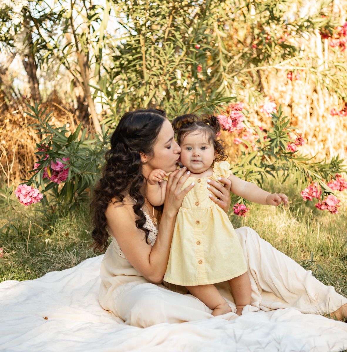 A tender outdoor portrait of a mother and child sitting on a white blanket surrounded by pink flowers and greenery.