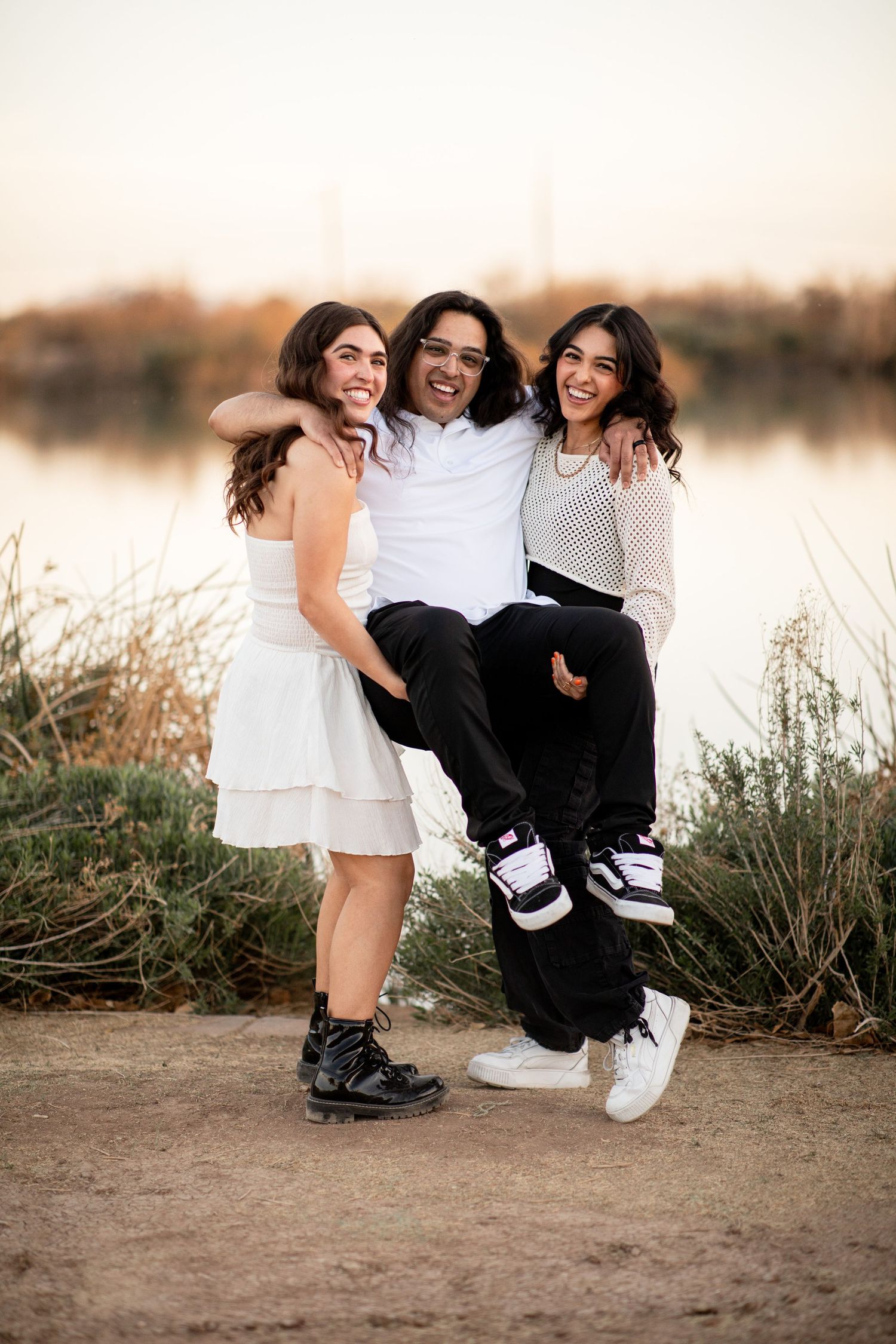 Three friends in matching black and white outfits pose casually together by a lake at sunset.