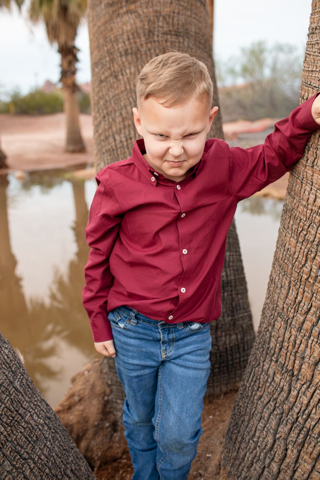 A young child in a burgundy shirt and jeans plays around a palm tree trunk near water in Papago Park in Phoenix, AZ.