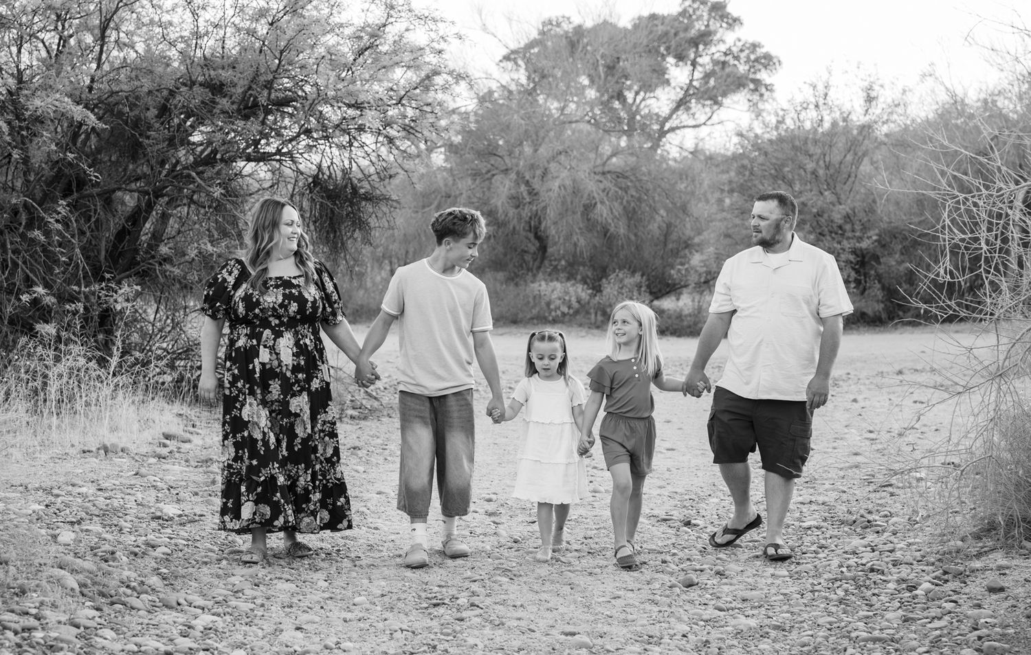 Black and white photo of a family of five holding hands while walking on a sandy path surrounded by trees.
