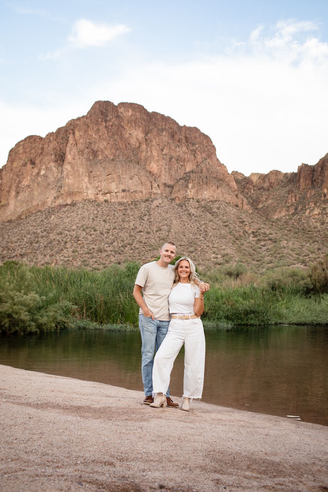 A couple stands together on a sandy beach with desert mountains and a serene lake in the background at Water Users recreational area.