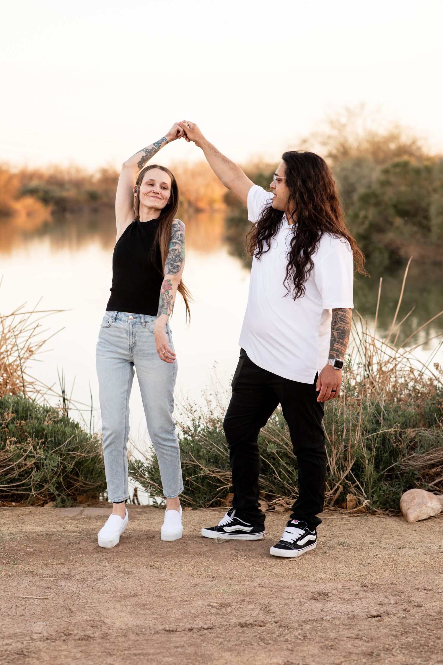 Two people dancing together by a lake at sunset, wearing casual outfits with tattoos visible and creating a joyful moment.