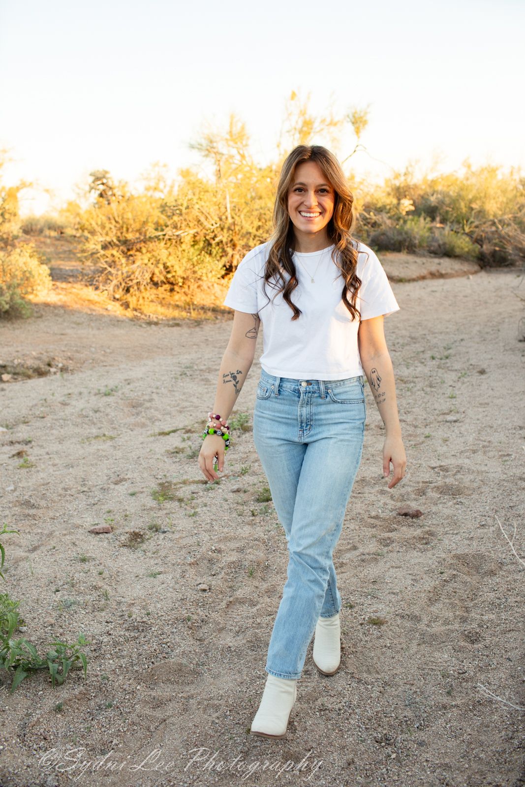 A casual outdoor portrait showing someone in a white t-shirt and light blue jeans walking on a desert path at sunset.