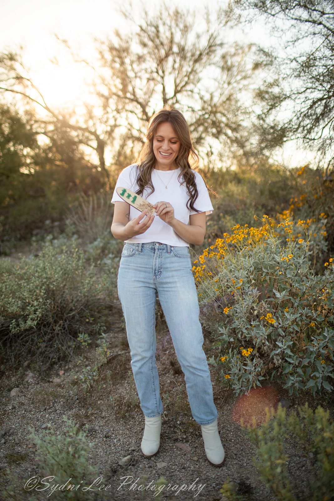 A person in a white t-shirt and light blue jeans poses among yellow wildflowers at sunset in a natural outdoor setting.