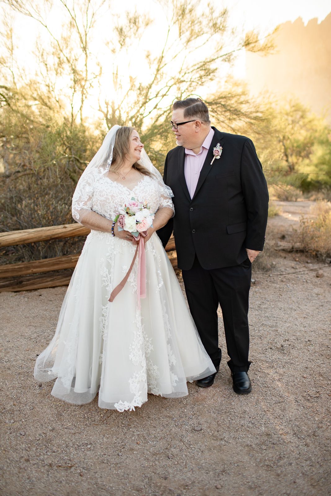 Wedding couple in formal attire pose together on a desert trail during golden hour.