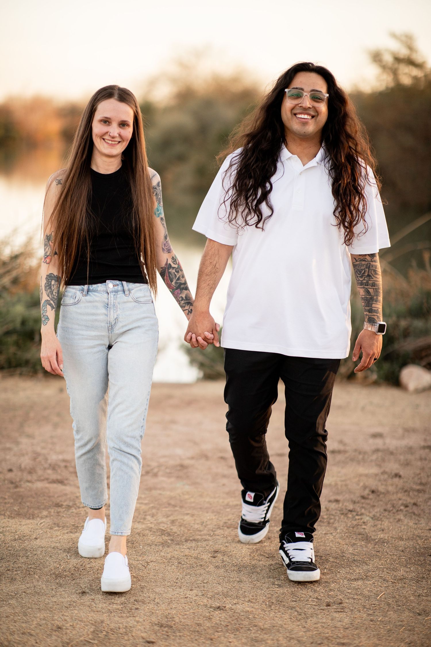 A couple holding hands while walking on a dirt path at sunset wearing casual outfits and sneakers.