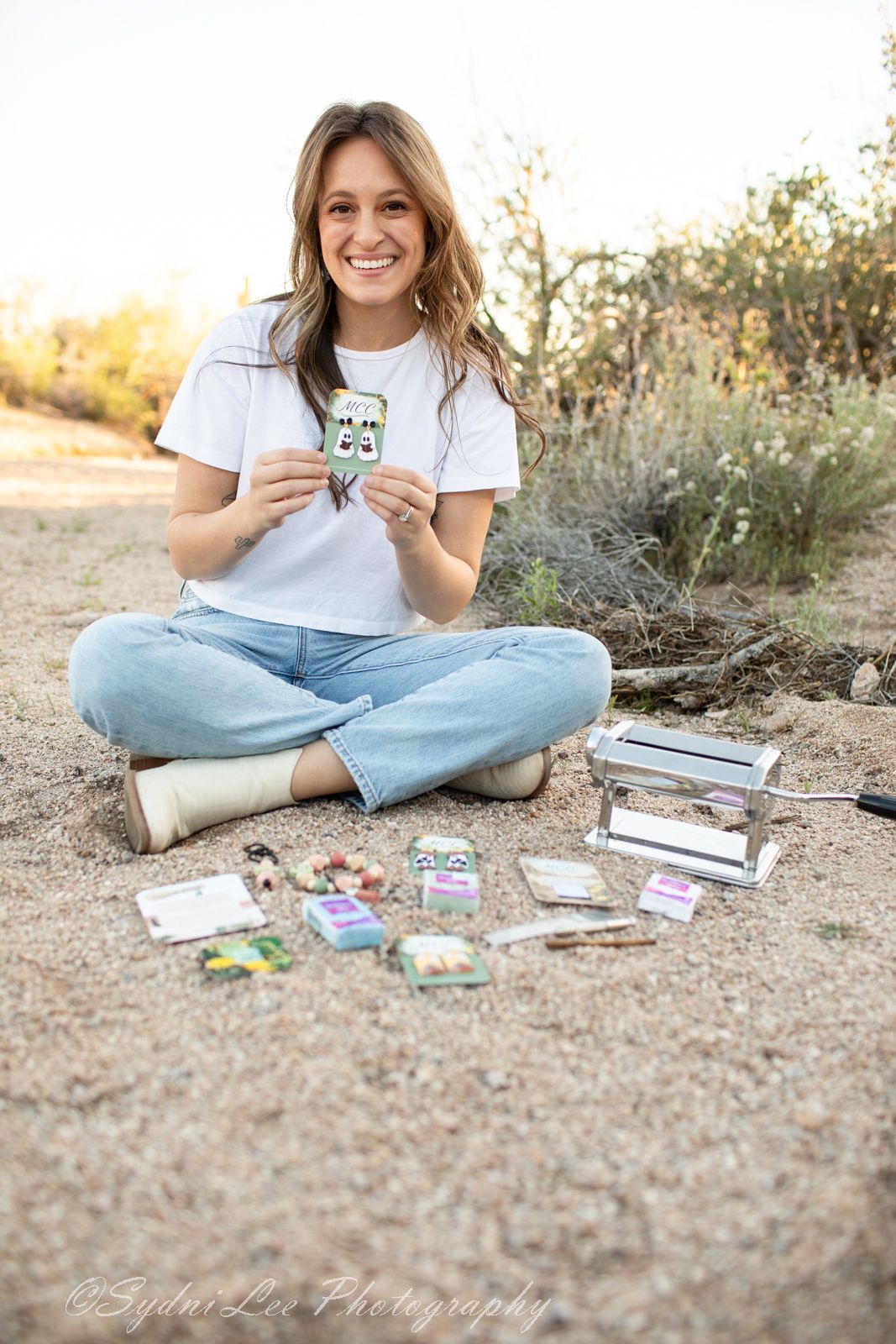 Person in white t-shirt and jeans sits cross-legged outdoors displaying small cards with wildflowers in background.