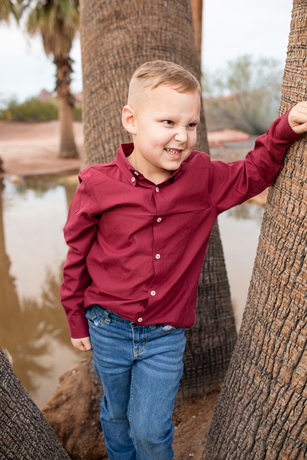 A child in a red button-down shirt and jeans poses playfully against a palm tree by the water in Papago Park in Phoenix, AZ.