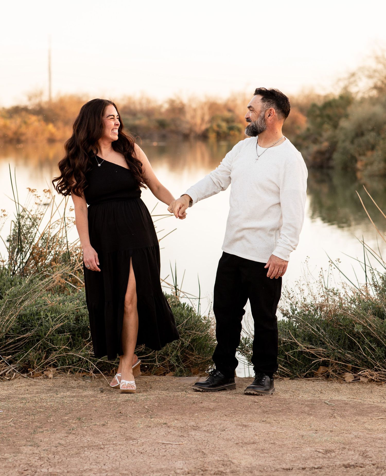 Couple holding hands during sunset photoshoot by a lake with tall grass and warm lighting.
