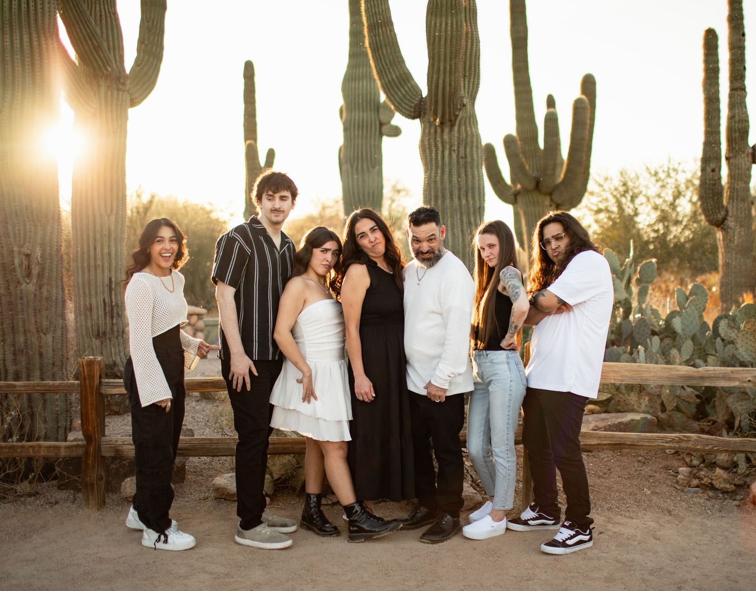 People in formal attire pose for photos in a desert setting with tall saguaro cacti and golden sunlight at sunset.