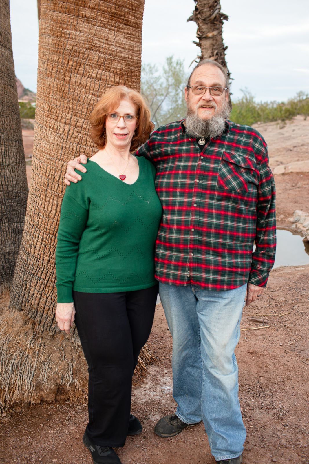 A couple poses together in front of palm trees, wearing casual winter clothing in Papago Park in Phoenix, AZ.