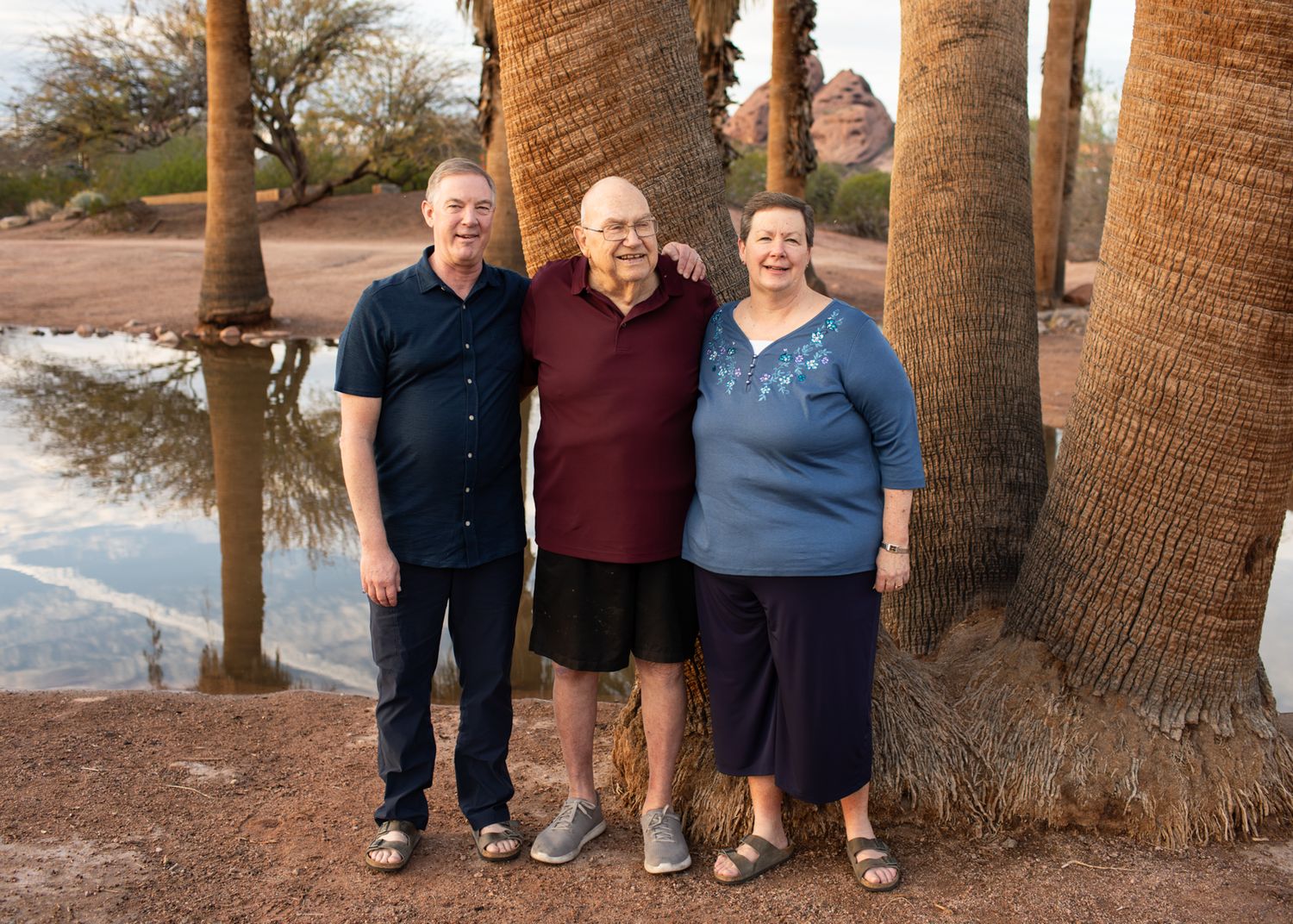 Three people standing together and smiling in front of tall palm trees near a body of water in Papago Park in Phoenix, AZ.