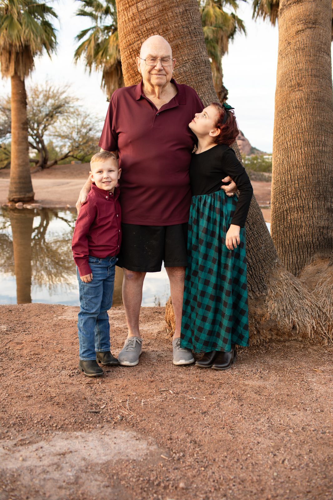 Family wearing burgundy and green plaid outfits poses together near palm trees at sunset in Papago Park in Phoenix, AZ.
