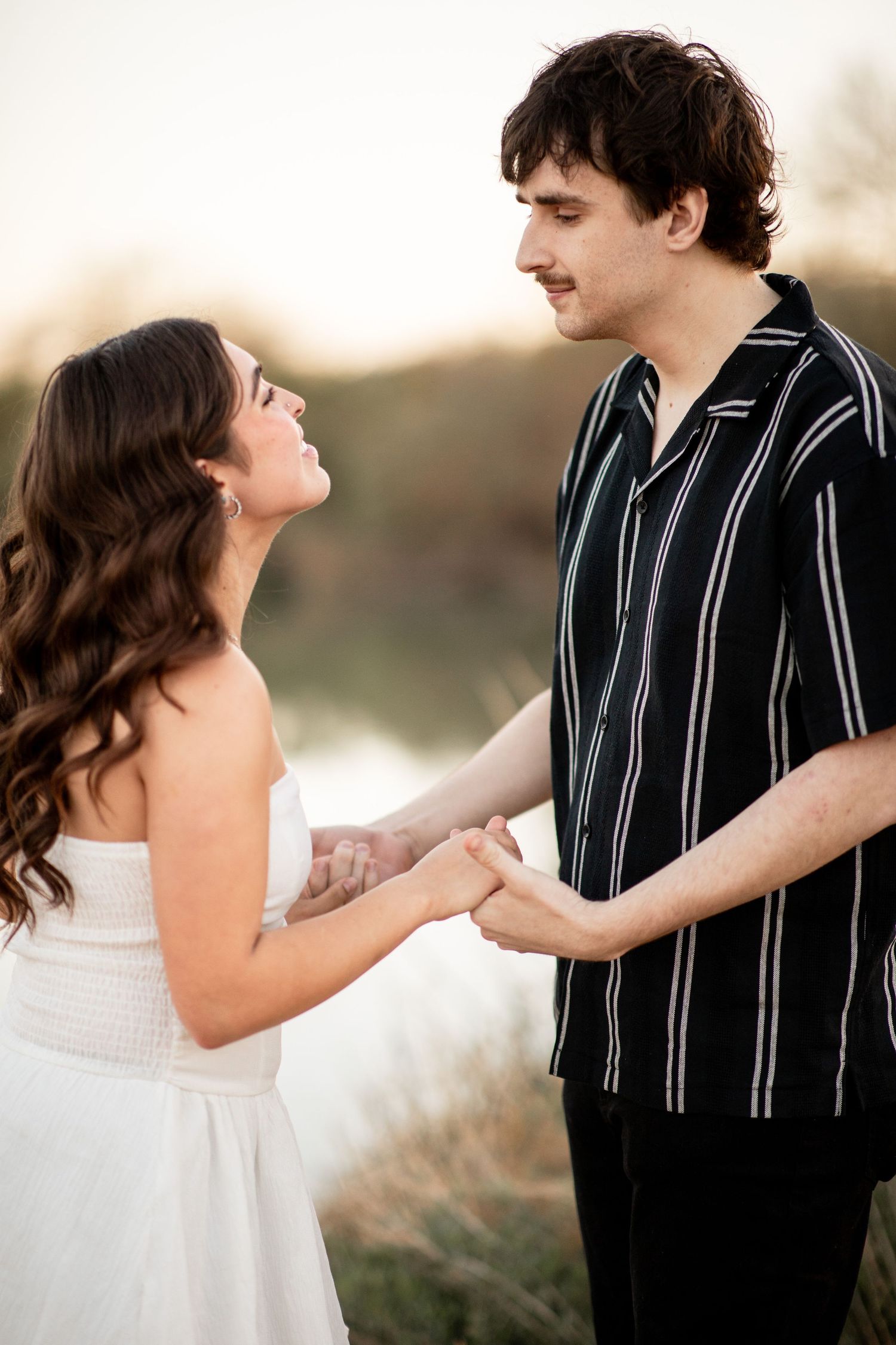A couple in wedding attire holds hands tenderly during a romantic outdoor photoshoot at sunset.