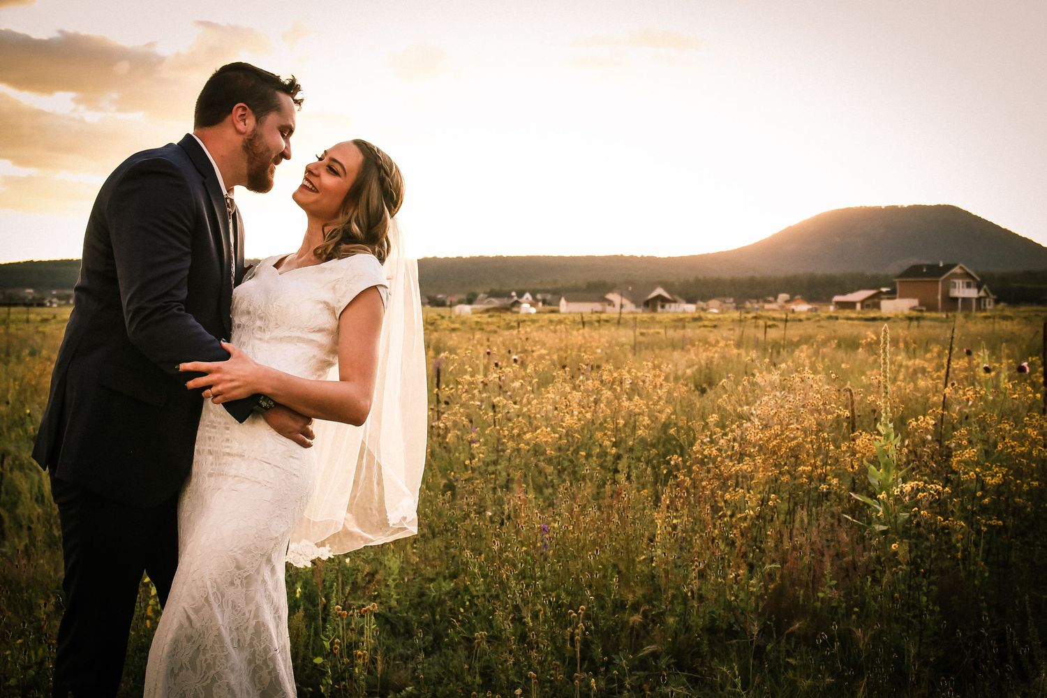 A couple in wedding attire embracing in golden field in Flagstaff at sunset with mountains in background.