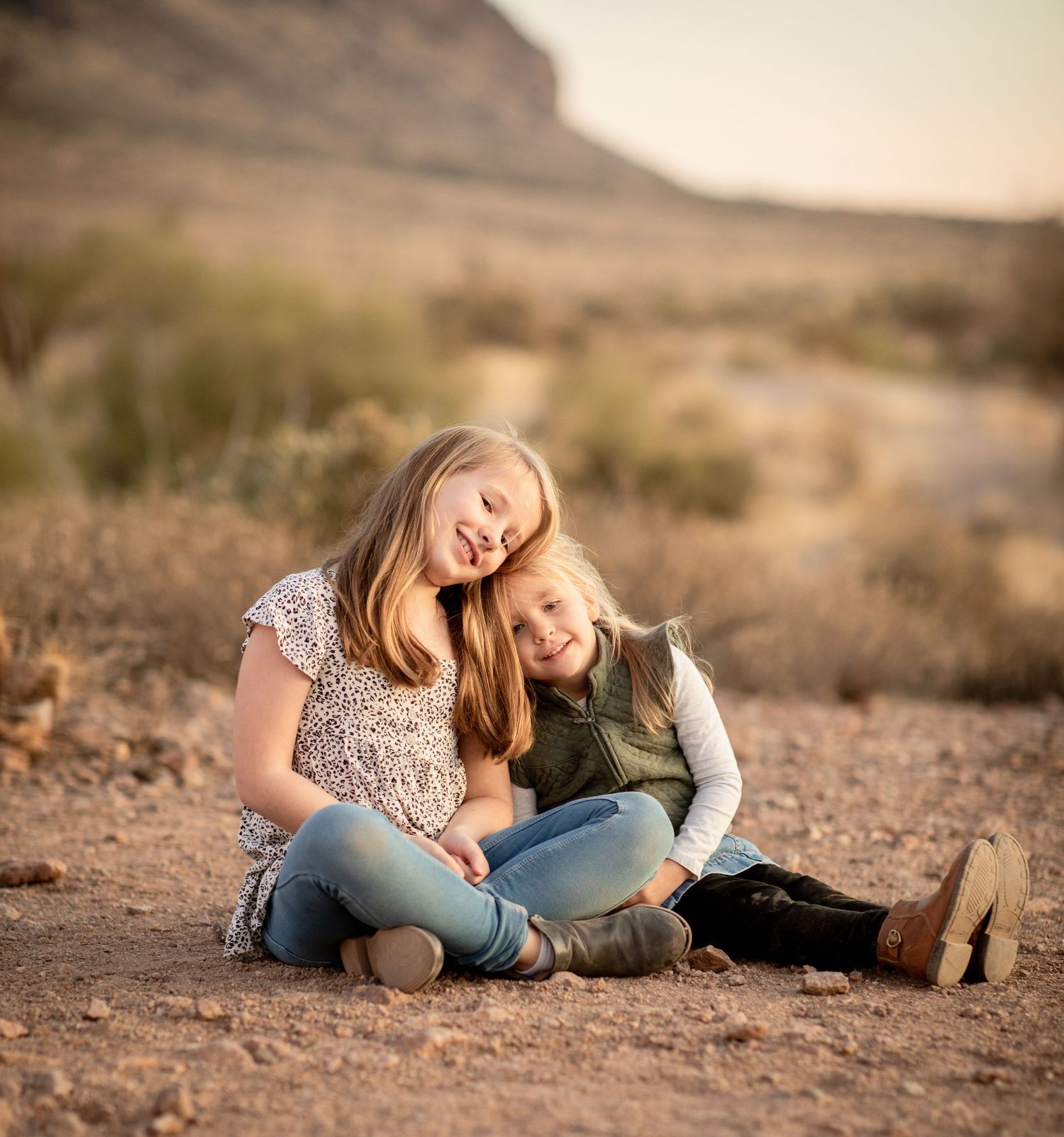 Two people sitting close together on desert ground with mountains and brush in background during golden hour at Lost Dutchman State park